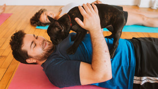 A man in a blue shirt and black shorts with white stripes on the side is laying down on a pink yoga mat. He is supporting a French Bulldog puppy who is standing on top of his chest with his two hands while smiling. In the background is a woman smiling.
